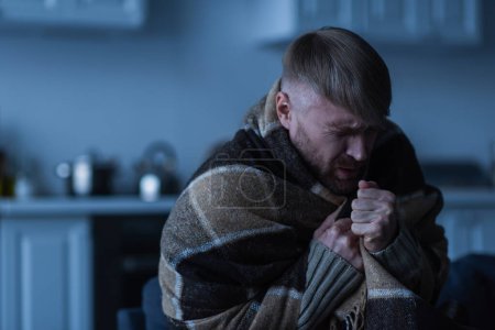 Gefrierender Mann mit geschlossenen Augen sitzt bei Stromausfall zu Hause in warmer Decke