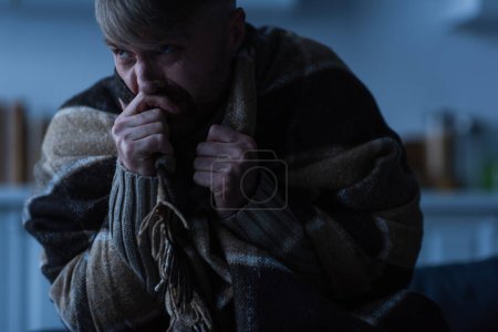 Photo for Stressed man holding hand near face while sitting under warm blanket and looking away in twilight - Royalty Free Image