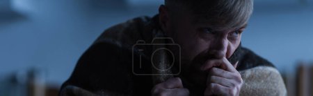 Photo for Depressed man sitting under warm blanket and holding hand near face during electricity shutdown, banner - Royalty Free Image