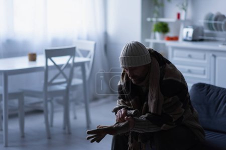 hombre congelado en sombrero de punto sentado en la cocina debajo de la manta y con guantes calientes