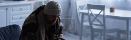 frozen man sitting in warm blanket and knitted hat at home in twilight, banner