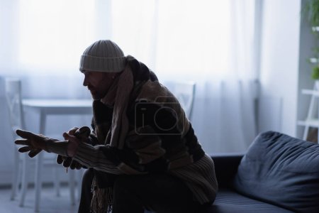 Photo for Side view of frozen man putting on gloves while sitting on couch in hat and warm blanket - Royalty Free Image