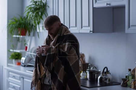 Gefrorener Mann steht in Küche unter warmer Decke und hält Tasse heißen Tee