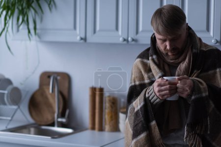 depressed and frozen man under warm blanket holding cup of hot tea in kitchen