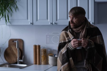 gefrorener Mann in warmer Decke hält Tasse mit heißem Getränk in der Küche und schaut weg