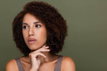 Joven mujer afroamericana tocando la barbilla y mirando hacia otro lado aislado en verde 