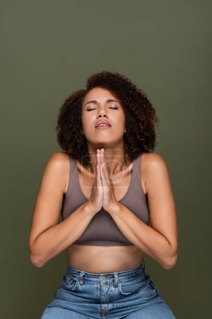Young african american woman in top with praying hands and closed eyes isolated on green 