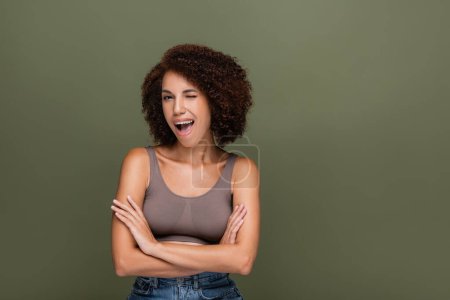 Pretty african american model crossing arms and winking at camera isolated on green 