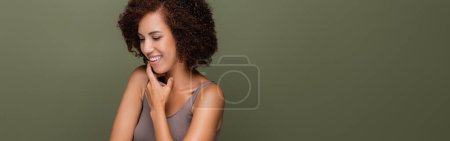 Jolie femme afro-américaine en haut touchant menton isolé sur vert, bannière 