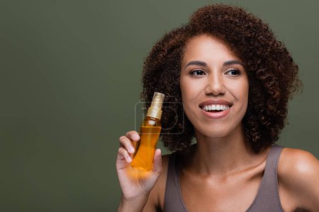 Curly african american woman holding cosmetic oil for hair isolated on green 