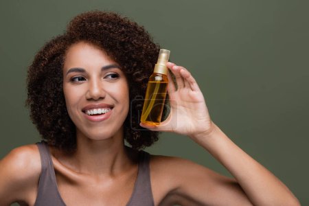 Lächelnde Afroamerikanerin betrachtet kosmetisches Haaröl isoliert auf grünem Grund 