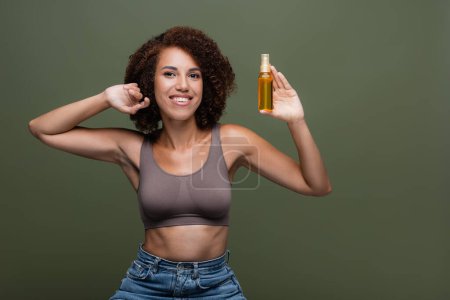 Curly african american woman in top and jeans holding cosmetic oil for hair isolated on green 