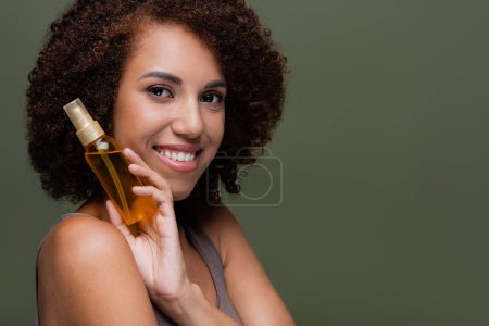 Porträt einer hübschen lockigen afrikanisch-amerikanischen Frau, die Kosmetiköl in der Hand hält und isoliert auf grün in die Kamera schaut 