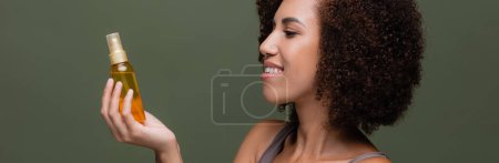 Téléchargez les photos : Vue latérale de la femme afro-américaine bouclée regardant l'huile cosmétique isolée sur vert, bannière - en image libre de droit