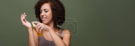 Femme afro-américaine joyeuse appliquant de l'huile cosmétique à la main isolée sur vert, bannière 