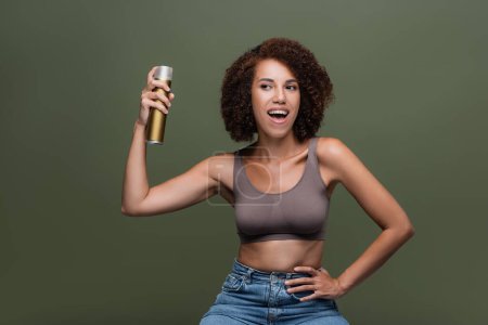 Cheerful african american woman in top holding hairspray isolated on green 