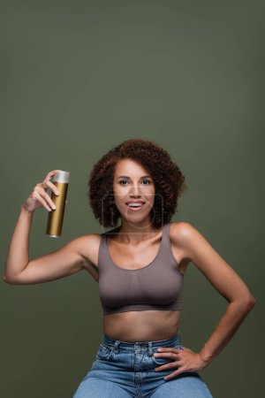 Photo for Pretty african american woman holding hairspray and looking at camera isolated on green - Royalty Free Image