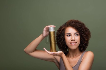 Photo for Smiling african american woman holding hairspray and looking away isolated on green - Royalty Free Image