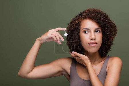African american woman in top holding cosmetic oil near curly hair isolated on green 