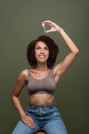 Positive african american woman in jeans and holding looking at bottle of cosmetic oil isolated on green 