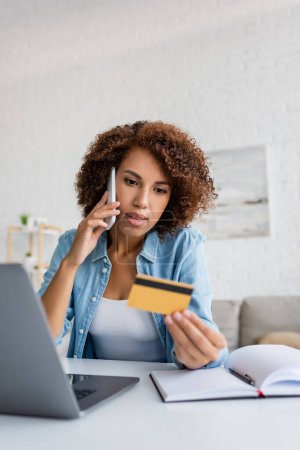 Mujer afroamericana hablando en smartphone y sosteniendo borrosa tarjeta de crédito cerca de portátil en casa 