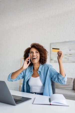 Young african american woman holding credit card and talking on smartphone at home 