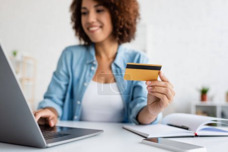 Verschwommene Afrikanerin hält Kreditkarte in der Hand und benutzt Laptop in der Nähe von Notizbuch auf Tisch 