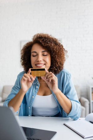 Pleased african american woman holding credit card near blurred laptop and notebook at home 