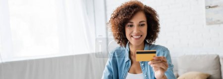 Smiling african american woman looking at credit card in living room, banner 