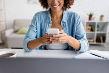 Vista recortada del alegre freelancer afroamericano utilizando un teléfono inteligente cerca de la computadora portátil y el té en casa 