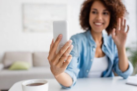 Téléchargez les photos : Femme afro-américaine floue ayant appel vidéo sur smartphone près de tasse de thé à la maison - en image libre de droit