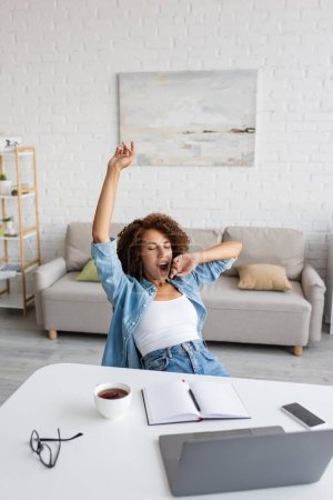 tired african american woman with closed eyes yawning and stretching near workplace at home
