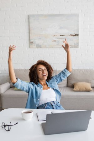 Téléchargez les photos : Femme afro-américaine joyeuse avec les yeux fermés se réjouissant près du lieu de travail à la maison - en image libre de droit