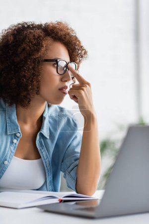 curly african american woman adjusting glasses and looking away near blurred laptop puzzle 620552916