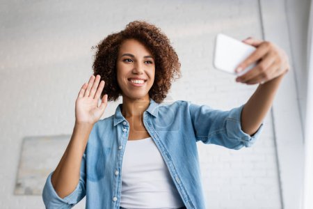 happy african american woman with curly hair waving hand during video call on smartphone