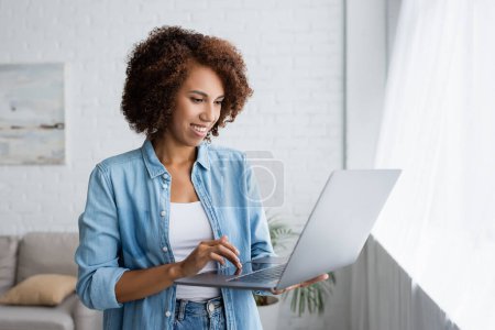 happy african american woman with curly hair holding laptop while working from home