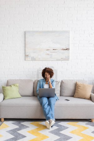 Foto de Alegre afroamericana mujer usando portátil mientras está sentado en el sofá y trabajando desde casa - Imagen libre de derechos