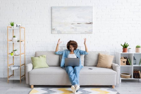 aufgeregte afrikanisch-amerikanische Frau benutzt Laptop, während sie auf der Couch sitzt und von zu Hause aus arbeitet 