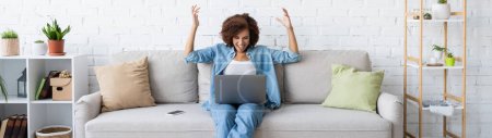 aufgeregte afrikanisch-amerikanische Frau mit Laptop, während sie auf der Couch sitzt und von zu Hause aus arbeitet, Banner