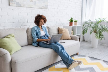 curly african american freelancer using laptop while sitting on couch and working from home 