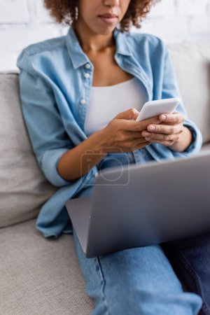 vista recortada de la mujer afroamericana mensajería en el teléfono inteligente cerca de la computadora portátil mientras está sentado en el sofá 