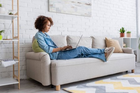 Foto de Mujer afroamericana enfocada usando el ordenador portátil mientras está sentado en el sofá y trabajando desde casa - Imagen libre de derechos