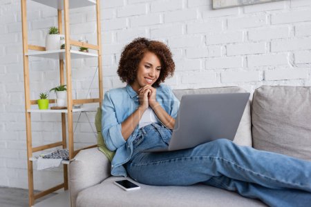 Téléchargez les photos : Heureuse femme afro-américaine assise avec les mains serrées et regardant un ordinateur portable près du smartphone sur le canapé - en image libre de droit