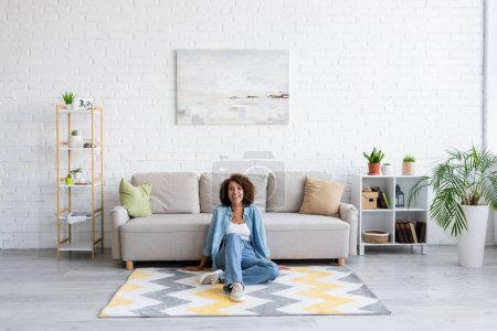 Foto de Alegre mujer afroamericana sentada en la alfombra con patrón cerca de sofá moderno en la sala de estar - Imagen libre de derechos