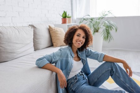 Foto de Mujer afroamericana positiva con el pelo rizado sentado cerca de sofá moderno en la sala de estar - Imagen libre de derechos