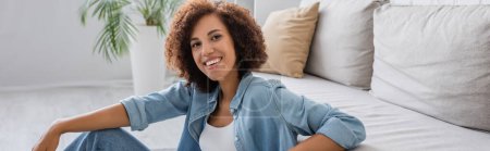 Foto de Mujer afroamericana positiva con el pelo rizado sentado cerca de sofá moderno en la sala de estar, pancarta - Imagen libre de derechos