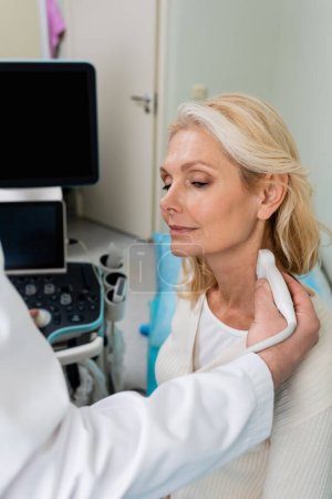 rubia de mediana edad mujer cerca de médico haciendo ultrasonido de su cuello en la clínica