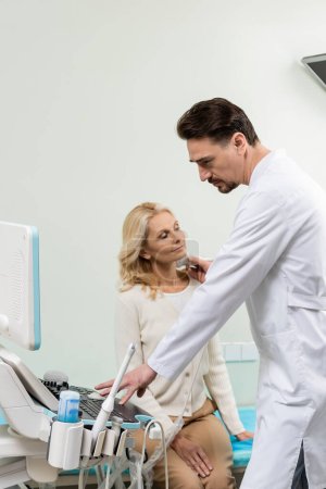 ernsthafter Arzt, der Ultraschallgerät bedient, während er die Diagnose der reifen Frau durchführt
