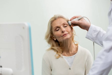 mujer rubia con los ojos cerrados cerca médico haciendo ultrasonido de cabeza