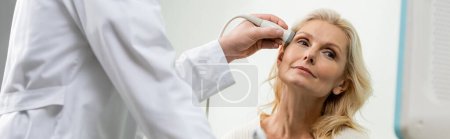 Blondine schaut in der Nähe von Arzt bei Ultraschalluntersuchung ihres Kopfes weg, Banner